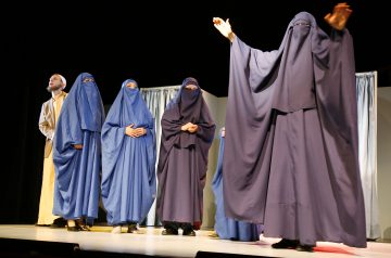 Lavalla seisoo viisi ihmistä, joista neljällä on sininen burka ja yhdellä ruskehtava puku. Yksi violettiin burkaan pukeutunut henkilö seisoo edessä kädet kohotettuna, kun taas muut seisovat rivissä takana yleisöön päin.