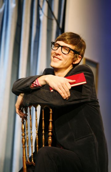 Hymyilevä henkilö, jolla on silmälasit ja tumma takki, istuu selkä menosuuntaan puisella tuolilla, kädet tuolin selkänojalla ja kädessään punainen kirja. Taustalla roikkuvat siniset verhot.