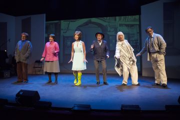 Kuusi värikkäisiin pukuihin pukeutunutta ihmistä seisoo rivissä lavalla ja esiintyy ilmeikkäästi ja ilmeikkäästi. Taustalla näkyy heijastettu kuva katukuvasta. Lavavalot valaisevat ryhmää.