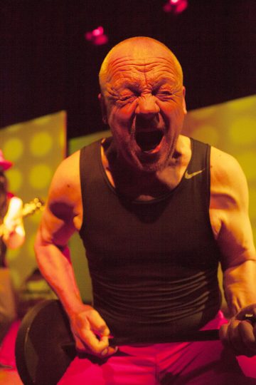 Kalju mies, jolla on musta toppi, laulaa tai huutaa intohimoisesti mikrofoniin lavalla, tarttuu mikrofonitelineeseen molemmin käsin, dramaattiset kasvonilmeet ja voimakas valaistus.