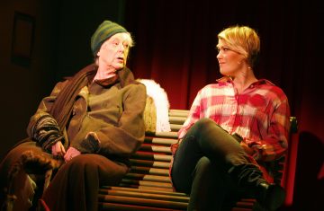 Kaksi ihmistä istuu penkillä näyttämövalaistuksen alla; toinen on pukeutunut nuhjuisiin talvivaatteisiin ja pipoon, kun taas toisella on punainen ruutupaita ja hän katsoo ensimmäistä henkilöä. Taustalla roikkuu punainen verho.