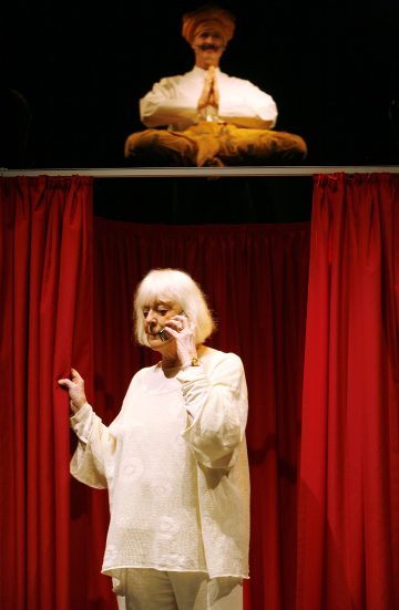 Valkoisiin hiuksiin pukeutunut vanhempi nainen seisoo punaisten verhojen edessä ja puhuu kuulokkeisiin. Hänen yläpuolellaan istuu turbaniin pukeutunut miespatsas tai hahmo, joka istuu ristissä kädet yhteen painettuina.