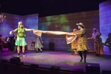 Lavalla on viisi värikkäisiin pukuihin pukeutunutta esiintyjää, joista kaksi etualalla nykii kultaista huivia. Tausta on valaistu eloisilla vihreillä ja violeteilla sävyillä. Lavalla vasemmalla puolella esiintyy pianisti.