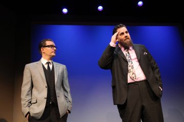 Kaksi pukumiestä seisoo lavalla, jossa on sininen tausta. Toinen mies, jolla on silmälasit ja harmaa puku, katsoo toista, jolla on parta, musta puku ja vaaleanpunainen paita ja joka tekee tervehdyseleen.