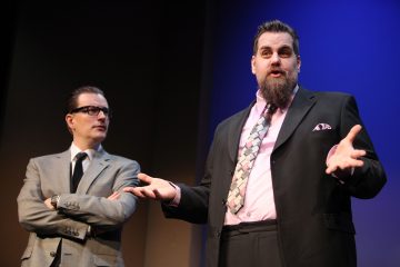 Kaksi pukumiestä seisoo lavalla kirkkaassa valaistuksessa. Vasemmalla olevalla miehellä on silmälasit ja hän ristii kätensä, kun taas oikealla olevalla miehellä on vaaleanpunainen paita ja kuviollinen solmio, ja hän puhuu eleitä molemmilla käsillään.