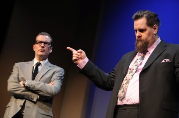 Lavalla seisoo kaksi pukumiestä. Vasemmalla oleva mies ristii kätensä ja hänellä on silmälasit, kun taas oikealla oleva mies osoittaa sormella ja hänellä on parta. Sininen ja ruskea valaistus luo dramaattisen tunnelman.