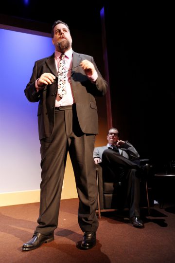 Pukuun ja solmioon pukeutunut mies seisoo ja puhuu lavalla kirkkaiden valojen alla, kun taas toinen pukuun pukeutunut mies istuu ja katselee taustalla olevalta tuolilta. Asetelma vaikuttaa muodolliselta ja ammattimaiselta.