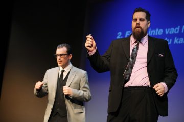 Kaksi pukumiestä seisoo lavalla; toisella on harmaa puku ja silmälasit, toisella musta puku ja vaaleanpunainen paita, ja heillä on kädessään esine, ja heidän takanaan on heijastettu sininen valkokangas.