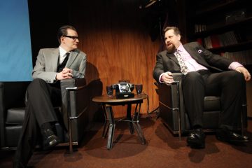 Kaksi pukumiestä istuu toisiaan vastapäätä nojatuoleissa, joiden välissä on pieni pöytä, jossa on musta pyörivä puhelin, huoneessa, jonka seinät on paneloitu puupaneelilla ja jossa on kirjahylly. Molemmat näyttävät keskustelevan.