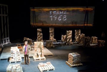 Lavaste, jossa on hajallaan tiiliä ja puisia kuormalavoja; niiden keskellä seisoo henkilö. Yläpuolella on valkokankaalla PRAHA 1968 valkoisin kirjaimin abstraktilla taustalla, mikä viittaa historialliseen tai dramaattiseen teatterikohtaukseen.
