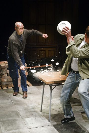 Vanhempi mies, jolla on silmälasit ja villapaita, heiluttaa mailaa ja rikkoo toisen miehen pitelemän lautasen, jonka sirpaleet ja roskat lentävät pöydälle.