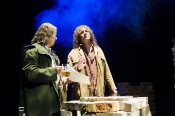 Kaksi miestä seisoo tiilipinon vieressä lavalla sinisten valojen alla; toisella on kädessään olut ja paperi, kun taas toinen elehtii dramaattisen esityksen aikana, jonka taustalla on savua.