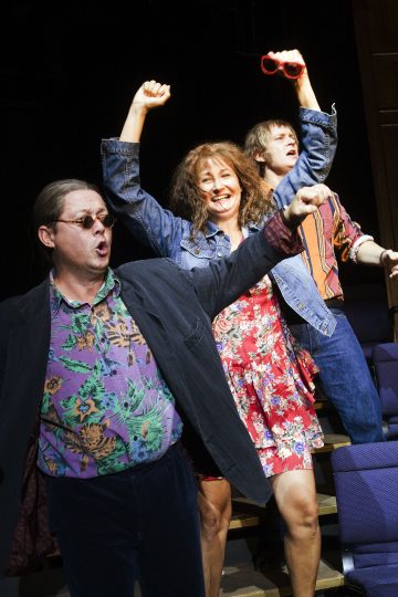 Kolme ihmistä laulaa tai hurraa innokkaasti teatterissa. Etummaisella henkilöllä on aurinkolasit, kukkapaita ja bleiseri. Takana punakukkaiseen kukkamekkoon pukeutunut nainen nostaa kädet ylös, ja toinen henkilö pitää punaisia aurinkolaseja päänsä yläpuolella.