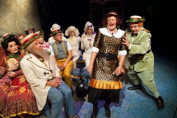 Lavalla esiintyy joukko värikkäisiin, omituisiin pukuihin pukeutuneita näyttelijöitä; yksi nainen seisoo ja laulaa tunteikkaasti, kun vihreäpukuinen mies elehtii dramaattisesti hänen vieressään, kun taas muut katselevat taustalla ilmeikkäin kasvoin.