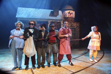 Kuusi näyttelijää värikkäissä, omituisissa puvuissa seisoo lavalla rivissä köyttä pitelemässä. Taustalla on lavaste, jossa on omituinen rakennus ja tornista ulos katsova ihminen, dramaattisessa teatterivalaistuksessa.