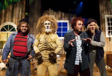 Lavalla seisoo neljä värikkäisiin pukuihin pukeutunutta näyttelijää; yksi on pukeutunut leijonaksi, ja muilla on raidalliset paidat ja hatut. He poseeraavat vilkkaasti ikkunoineen ja huonekaluineen varustetun puisen lavastuksen edessä näyttämövalaistuksen alla.