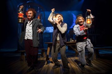 Kolme värikkäisiin, vanhanaikaisiin pukuihin pukeutunutta näyttelijää pitelee lyhtyjä ja esiintyy tarmokkaasti hämärästi valaistulla näyttämöllä, jossa on merellinen tausta, mikä viittaa vilkkaaseen teatteriesitykseen.