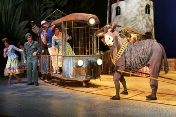 Värikkäisiin pukuihin pukeutuneet näyttelijät esiintyvät lavalla; jotkut ajavat omituisella raitiovaunulla, ja aasin pukuun pukeutunut henkilö vetää sitä, ja taustalla on eläväinen, pyöreä, tornimainen rakennus.