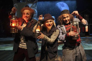 Kolme näyttelijää, jotka ovat pukeutuneet omituisiksi merimiehiksi, joilla on sotkuinen tukka ja puvut, pitävät lyhtyjä näyttämöllä ja hymyilevät dramaattisessa valaistuksessa, ja taustalla on kuun valaisema merimaisema.