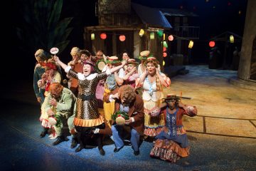 Ryhmä värikkäisiin, omituisiin pukuihin pukeutuneita näyttelijöitä esiintyy tarmokkaasti lavalla, hattuja ja rekvisiittaa kädessään, ja taustalla on juhlallisia lyhtyjä ja maalaismainen lavastus.