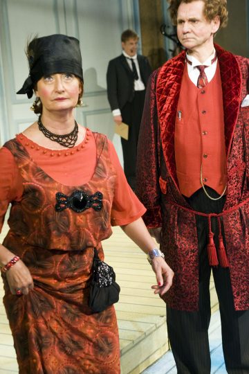 Punakuvioiseen mekkoon ja mustaan huiviin pukeutunut nainen seisoo punaliiviin ja kaapuun pukeutuneen miehen vieressä. Molemmilla on vakavat ilmeet. Taustalla seisoo smokkiin pukeutunut mies sumeana lavasteessa.