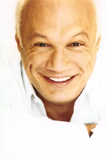 Hymyilevä vanhempi mies, jolla on lyhyet vaaleat hiukset, on pukeutunut valkoiseen paitaan ja katsoo suoraan kameraan valkoista taustaa vasten.