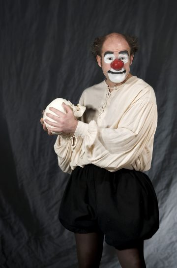 Klovniksi pukeutunut henkilö, jolla on valkoinen kasvomaali, punainen nenä ja vaalea paita, pitää kädessään ihmisen kalloa ja seisoo tumman, ryppyisen taustan edessä.