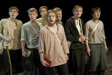 Seitsemän yksinkertaisiin, vanhanaikaisiin vaatteisiin pukeutunutta nuorta miestä seisoo lähekkäin näyttämöllä ja katsoo vakavin tai huolestunein ilmein eteenpäin, mikä viittaa teatteriesityksen dramaattiseen hetkeen.