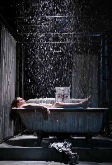 Henkilö makaa vanhanaikaisessa kylpyammeessa rankkasateessa sisätiloissa, dramaattinen valaistus ja osittain suljettu suihkuverho taustalla. Vesi roiskuu ja virtaa alas, mikä luo dramaattisen tunnelman.