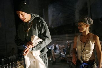 Kaksi ihmistä seisoo ostoskärryjen vieressä hämärässä. Toisella on pipo, kultaketju ja verkkarit, ja hänellä on valkoinen kassi kädessään. Toinen, jolla on harmaa lippis ja toppi, katsoo hymyillen kohti ensimmäistä henkilöä.