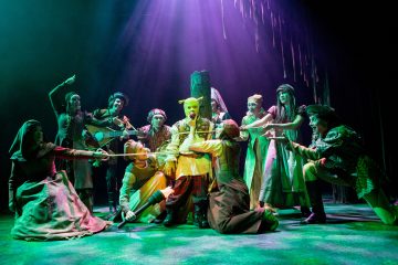 Näyttämökohtaus, jossa keskiaikaisiin pukuihin pukeutuneet näyttelijät ympäröivät pylvääseen sidottua Shrekiksi pukeutunutta hahmoa dramaattisessa vihreässä ja violetissa valaistuksessa. Näyttelijät osoittavat Shrekiä ilmeikkäin elein.