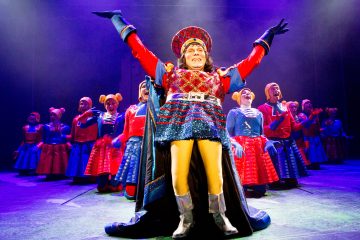 Värikkääseen, taidokkaaseen pukuun pukeutunut esiintyjä seisoo näyttämön keskellä kädet kohotettuna, ja häntä ympäröivät tanssijat, joilla on samansävyiset punaiset ja siniset asut, kirkkaassa näyttämövalaistuksessa.