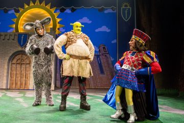 Lavalla esiintyy kolme näyttelijää, joista yksi on aasi, yksi Shrek, jolla on vihreä iho ja pehmustettu vatsa, ja yksi lordi Farquaad punasinisessä asussa, linnan taustan ja kirkkaan auringon edessä.