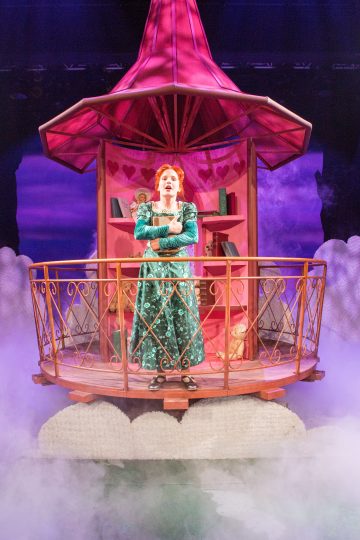 Pitkään vihreään mekkoon pukeutunut esiintyjä seisoo sydämillä ja kirjoilla koristellulla pyöreällä parvekkeella pilvien ja vaaleanpunaisen valaistuksen ympäröimänä, ja taustalla on omituinen, satukirjamainen tausta.