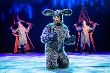 Harmaaseen aasipukuun pukeutunut esiintyjä polvistuu näyttämön keskellä, ja kaksi muuta ritareiksi pukeutunutta esiintyjää pitelee punaisia rekvisiittaa taustalla sinisessä näyttämövalaistuksessa.