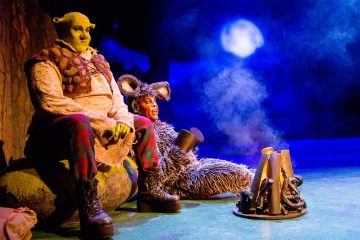 Kaksi pukuihin pukeutunutta näyttelijää, joista toinen on Shrek ja toinen Aasi, istuvat lavalla sinisellä taustavalaistulla ja täysikuun valossa tekaistun leirinuotion äärellä ja esittävät kohtauksen Shrek-musikaalista.