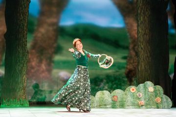 Lavalla tanssii nainen, jolla on vihreä kukkamekko ja kruunu, ja hänellä on kukkakori kätensä ojennettuna. Tausta muistuttaa lumottua metsää, jossa on maalattuja puita ja pensaita.