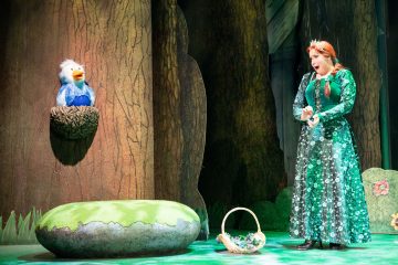 Vihreään, kimaltelevaan mekkoon pukeutunut nainen seisoo lavalla korin ja suuren kiven vieressä ja laulaa tai puhuu yllättynyt ilme kasvoillaan, kun sininen lintunukke istuu puuhun kiinnitetyssä pesässä.