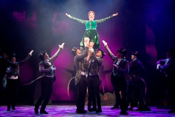 Joukko pukuihin pukeutuneita esiintyjiä nostaa vihreään mekkoon pukeutuneen naisen päänsä yläpuolelle lavalla, jossa on violetti valaistus ja metsätausta. Nainen hymyilee kädet kohotettuna, kun muut katsovat ylös ja kurottelevat häntä kohti.