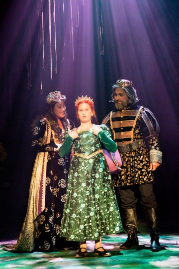 Kolme keskiaikaiseen pukuun pukeutunutta ihmistä seisoo violettien näyttämövalojen alla. Keskellä seisoo nuori henkilö vihreässä mekossa ja kruunussa, ja häntä reunustaa kaksi aikuista, joilla on kuninkaalliset asut ja kruunut, luoden dramaattisen teatterikohtauksen.