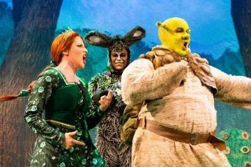 Lavalla esiintyy kolme värikkäisiin pukuihin pukeutunutta näyttelijää, joista yksi on beigeen pukeutunut vihreä örkki, toinen vihreäpukuinen nainen ja kolmas aasin hahmo metsätaustaa vasten. He näyttävät laulavan tai puhuvan.