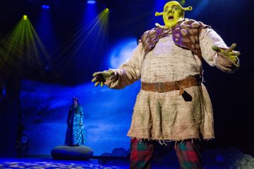 Shrekiksi pukeutunut esiintyjä, jolla on vihreä iho ja jättikorvat, seisoo lavalla dramaattisessa valaistuksessa. Taustalla näkyy toinen hahmo seisomassa kalliolla kuun valaiseman taustan edessä.