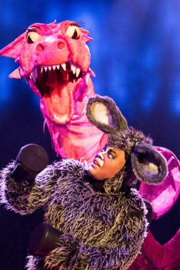 Lavalla seisoo esiintyjä, jolla on pörröinen harmaa aasipuku, jossa on suuret korvat, ja katselee suurta vaaleanpunaista lohikäärme-nukkea, jolla on terävät hampaat ja suuret silmät sinistä taustaa vasten.