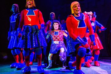 Ryhmä värikkäisiin, futuristisiin pukuihin pukeutuneita näyttelijöitä esiintyy lavalla. Useimmilla on punaiset paidat, siniset hameet ja keltaiset kypärät, kun taas yksi keskellä on pukeutunut sinipunaiseen asuun ja polvistuu. Valaistus on dramaattinen ja eläväinen.