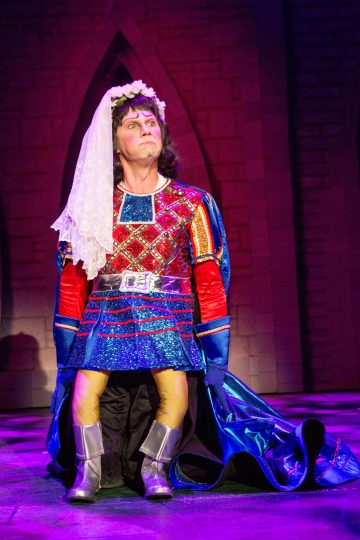 Kirkkaaseen, värikkääseen keskiaikaiseen pukuun, huntuun, viittaan ja hopeisiin saappaisiin pukeutunut henkilö polvistuu näyttämöllä dramaattisessa valaistuksessa kivimäisen taustan edessä.