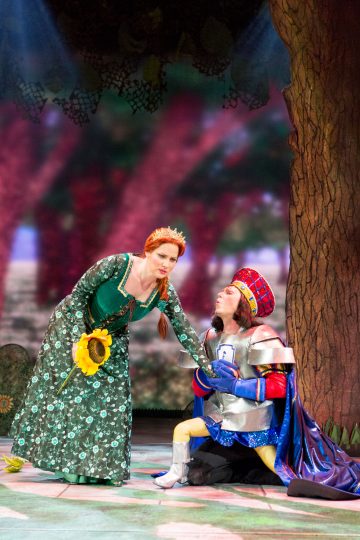 Vihreään mekkoon ja kruunuun pukeutunut nainen pitelee auringonkukkaa kumartuen kohti miestä, jolla on kiiltävä haarniskapuku ja punaruutuinen kypärä, joka polvistuu näyttämöllä ja kurottautuu kädelle teatterimaisessa ulkoilmaympäristössä.