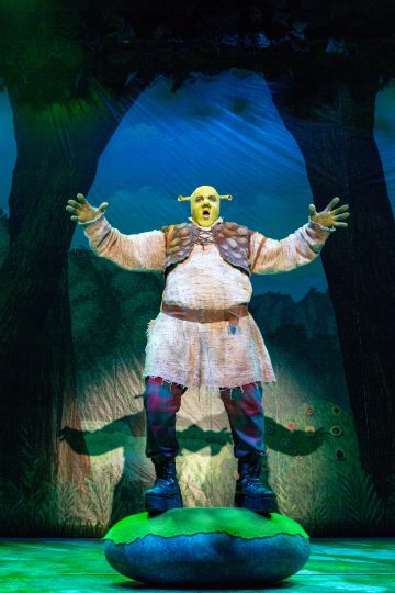 Shrekiksi pukeutunut esiintyjä, jolla on vihreä meikki, korvaproteesit ja puku, seisoo lavalla, jossa on metsämaisemia, kädet kohotettuna ja suu auki ikään kuin laulaisi tai huutaisi.