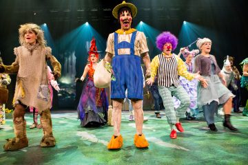 Ryhmä värikkäisiin, omituisiin pukuihin pukeutuneita näyttelijöitä esiintyy energisesti lavalla, ja yksi klovnin näköinen hahmo keltaisissa kengissä ja sinisessä asussa seisoo kirkkaiden valojen keskellä.