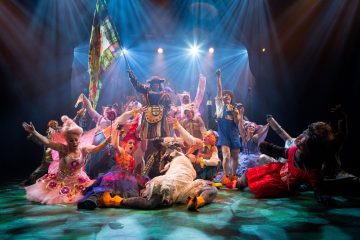 Värikkäisiin, omituisiin pukuihin pukeutunut vilkas joukko esiintyjiä poseeraa energisesti lavalla kirkkaiden valonheittimien alla ja herättää teatterituotannossa jännityksen ja juhlan tunteen.