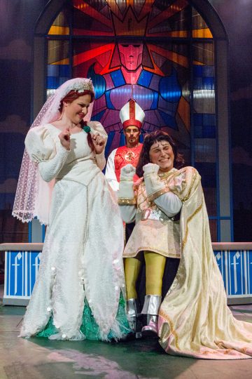 Kolme näyttelijää esiintyy lavalla komediallisessa hääkohtauksessa: morsian valkoisessa mekossa, kullanhohtoiseen kaapuun pukeutunut henkilö istuu ja hymyilee, ja taustalla seisoo piispa punavalkoisissa kaavuissa.